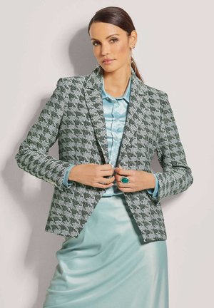 Grüner und weiß gemusterter Blazer mit scharfen Revers, kombiniert mit einer hellblauen Satinbluse und einem Rock. Auffällige Akzente setzen ein großer türkisfarbener Ring.