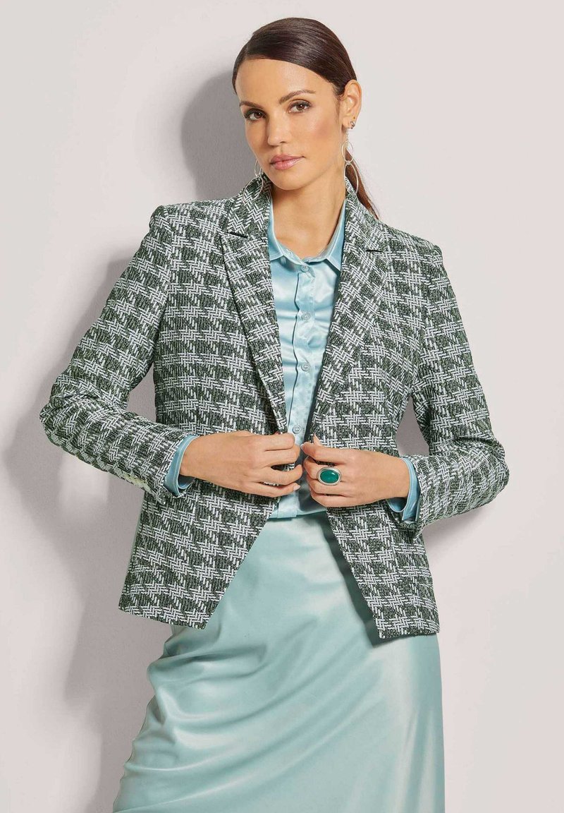 Grüner und weiß gemusterter Blazer mit scharfen Revers, kombiniert mit einer hellblauen Satinbluse und einem Rock. Auffällige Akzente setzen ein großer türkisfarbener Ring.