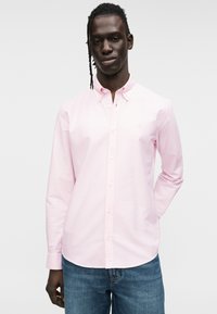 Chemise à manches longues rose clair en coton, dotée d'un col et d'un logo discret sur la poitrine. Associée à un jean en denim foncé.