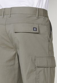 Primo piano di pantaloni cargo verde oliva con tasche a patta ai lati e sul retro, indossati con una camicia bianca.
