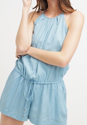 Mujer que lleva un mono sin mangas de estampado azul con cuello halter y cintura ajustable con cordón, de pie con un brazo cruzado y el otro levantado cerca de su rostro.
