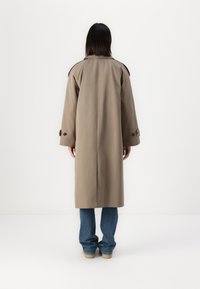 Abrigo trench de color caqui con corte recto, mangas anchas y acentos de botones. El abrigo cuenta con una abertura en la espalda y se combina con jeans azules.