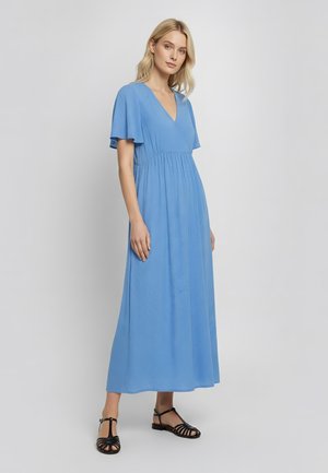 Vrouw met blond haar, gekleed in een lichtblauwe maxi-jurk met V-hals en vliegermouwen, en zwarte sandalen met bandjes en platte zolen, staand tegen een effen achtergrond.