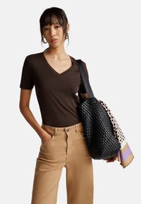 Vrouw in een bruine V-hals shirt en beige broek, met een zwarte geweven schoudertas waarover een patroon sjaal is gedrapeerd.