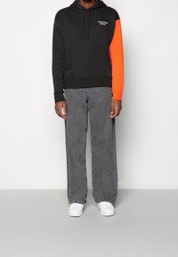 Sudadera negra con una manga naranja, con un bolsillo frontal y un logo. Combinada con unos vaqueros grises holgados y zapatillas blancas.