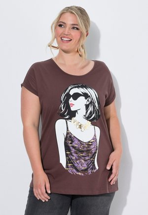 Smiling woman with blonde hair wears brown T-shirt featuring graphic of a woman with sunglasses and a purple patterned top.