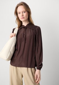 Brun langærmet bluse med en rynket krave, bindebånd ved halsen og strukturerede skuldre. Sammen med beige bukser og en hvid taske.