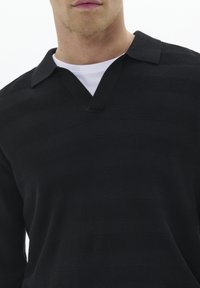 Camisa polo negra texturizada con un profundo escote en V, que presenta un patrón de canalé y mangas cortas, superpuesta sobre una camiseta blanca.