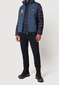 Giacca puffer blu navy con frontale in pile blu, colletto alto e toppe. Abbinata a pantaloni della tuta scuri e stivali neri con accenti rossi.