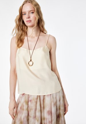 Mujer rubia con una camiseta de tirantes crema, un collar largo con colgante y una falda plisada en tonos rosa y beige.