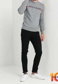 Sudadera gris con el texto "HILFIGER" y una franja roja/blanca a lo largo del pecho, combinada con jeans skinny negros y zapatillas blancas.