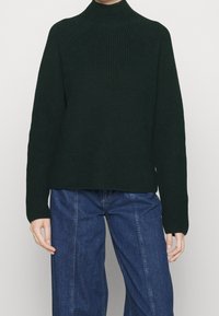 Donkergroen geribde coltrui met een ontspannen pasvorm en lange mouwen. Het materiaal lijkt textuur en gebreid te zijn. Gepaard met lichtblauwe wijde jeans.