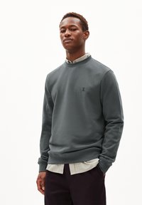 Sweatshirt gris avec un col arrondi, des poignets et un ourlet côtelés. Présente un petit logo sur la poitrine. Porté par-dessus une chemise à carreaux légère.