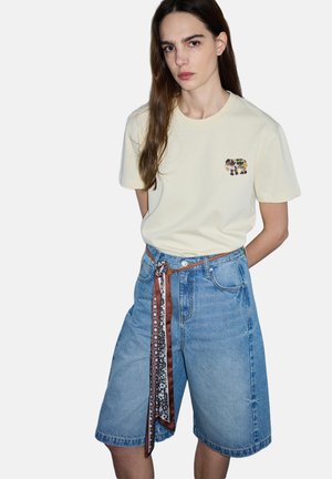 Frau trägt cremefarbenes T-Shirt mit kleinem Motiv und lockere, mittellange Denim-Shorts, die mit braunen und gemusterten Seidenschals gebunden sind.