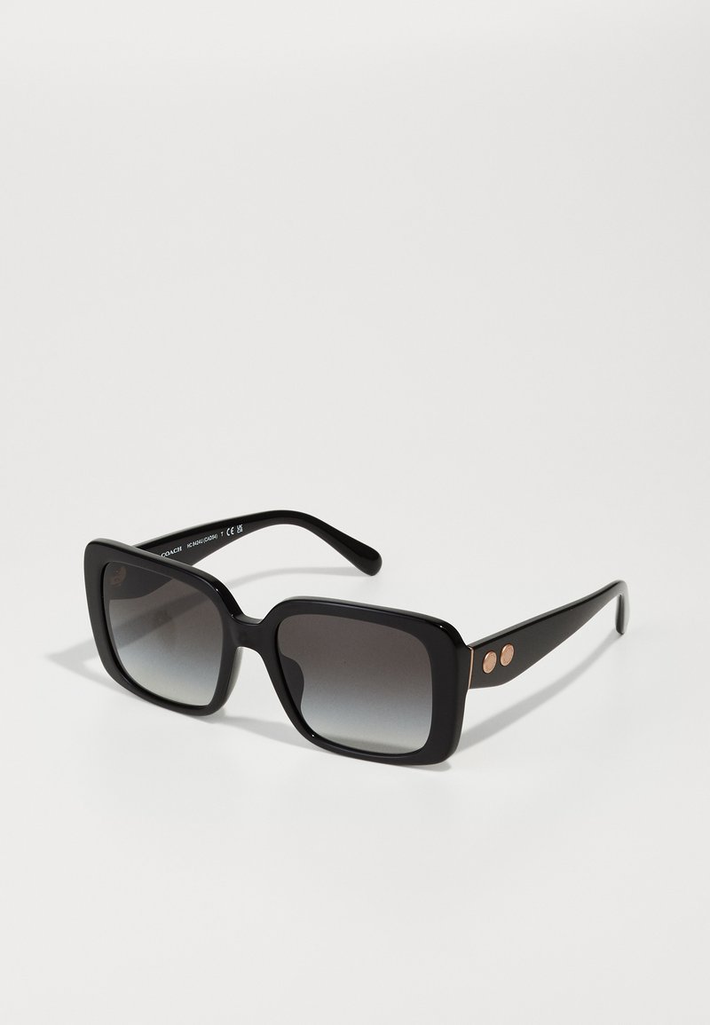 Lunettes de soleil noires oversize avec une monture carrée, des verres gris dégradés et deux accents dorés sur les branches. Texture plastique lisse.