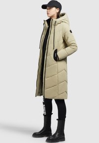 Gaišs bežs pūšammais mētelis ar kapuci un melniem akcentiem, ar izšūta raksta, ar rāvējslēdzēju priekšā un sānu kabatām. Nēsāts ar melnām legingām un zābakiem.