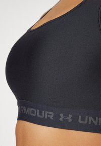 Svart sportsbh med glatt stoff, racerback-design og en bred elastisk band preget med ordet "ARMOUR" i grått.