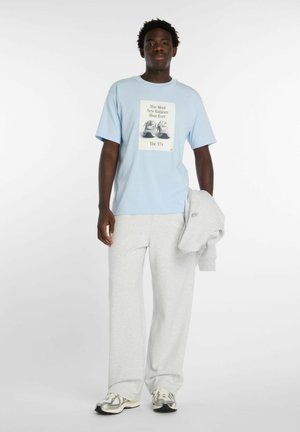 Mann trägt hellblaues New Balance Grafik-T-Shirt, hellgraue Jogginghose und Turnschuhe, hält ein gefaltetes graues Sweatshirt.