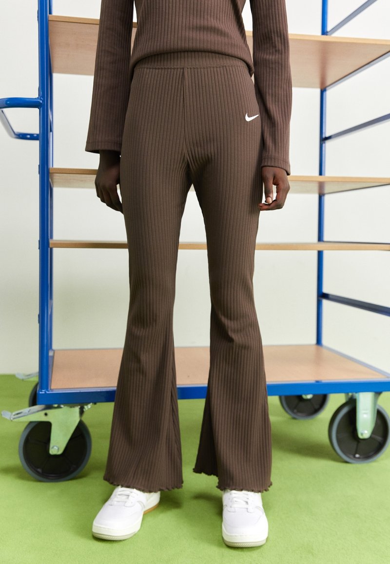 Bruna ribbade utsvängda byxor med hög midja och vitt Nike-logotyp. Materialet har en mjuk textur; fållen är kantad med en vågig kant.