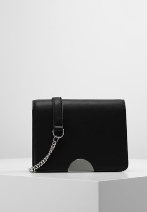 Schwarze rechteckige Clutch mit silberner Kettenriemen und halbkreisförmigem silbernem Metallschmuck vorne, auf weißem Block platziert.