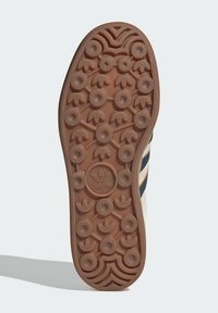 Rubber outsole with a textured tan surface, featuring circular and geometric patterns. Centered logo displays the brand name.