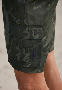 Maskovacie cargo šortky v tmavozelenej látke, s viacerými vreckami, prešitým lemom a značkou na boku s číselnými detailmi.