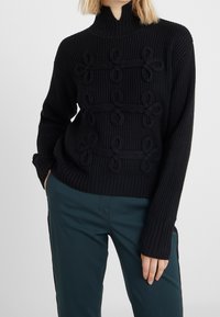 Pull noir côtelé à col montant, présentant des motifs de boucles texturées sur le devant. Manches longues, assorti à un pantalon bleu sarcelle.