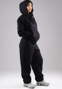 Woman in black hoodie and sweatpants with hands in pockets, standing in profile wearing silver athletic shoes against a plain background.