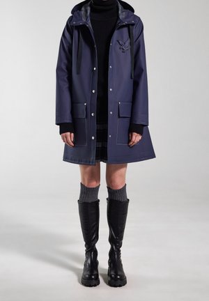 Personne portant un imperméable bleu marine à capuche, un pull col roulé noir, une jupe à carreaux, des chaussettes grises montantes et des bottes noires hautes jusqu'aux genoux, debout sur un fond gris.