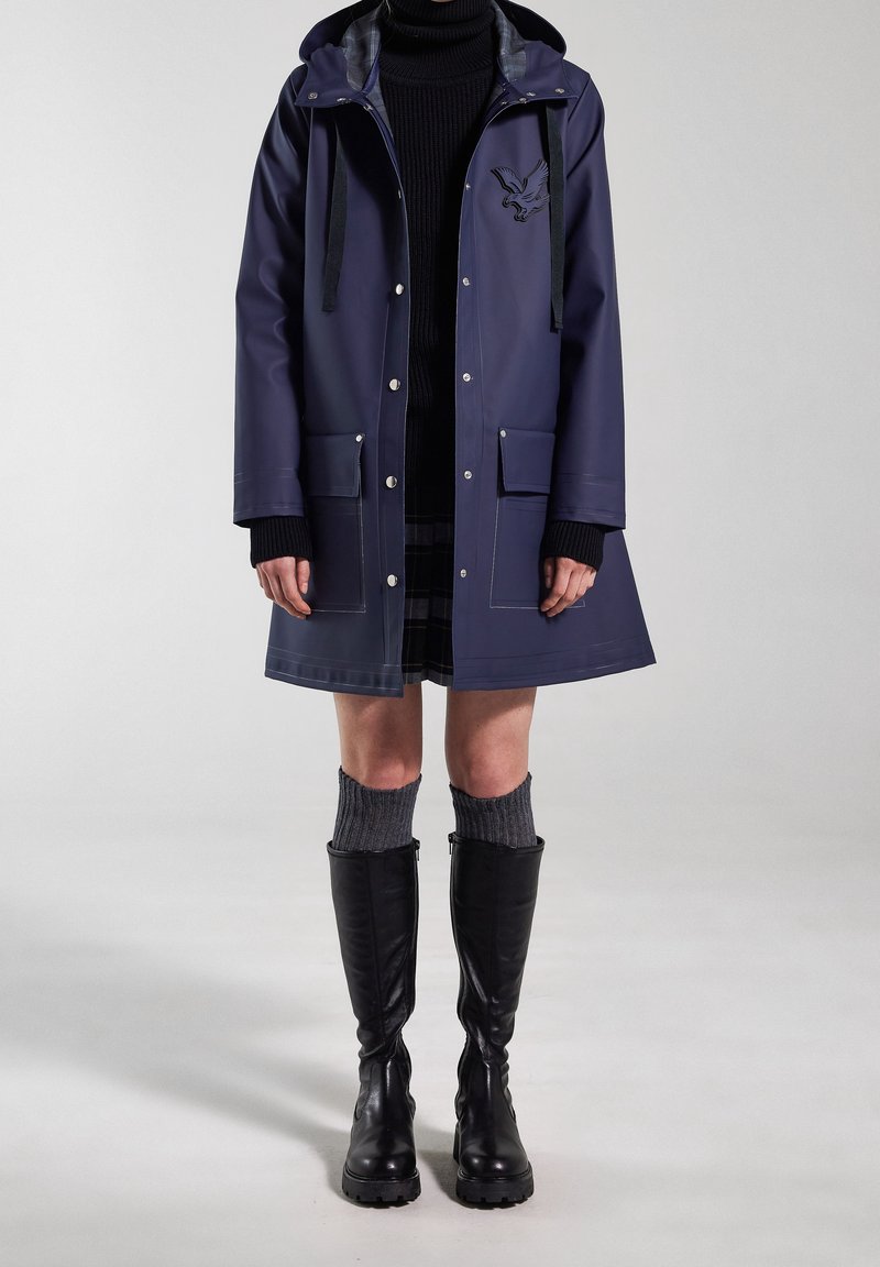 Personne portant un imperméable bleu marine à capuche, un pull col roulé noir, une jupe à carreaux, des chaussettes grises montantes et des bottes noires hautes jusqu'aux genoux, debout sur un fond gris.