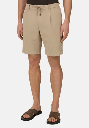 Beige Herren-Shorts in knieumspielender Länge mit Kordelzugbund, Seitentaschen und braunen Sandalen mit Kreuzriemen an hellhäutigen Beinen.