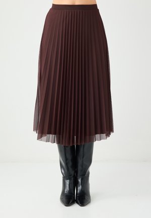 Brauner plissierter Rock aus durchsichtigem Stoff, geschichtet mit einem blickdichten Unterrock, kombiniert mit kniehohen schwarzen spitzen Stiefeln.