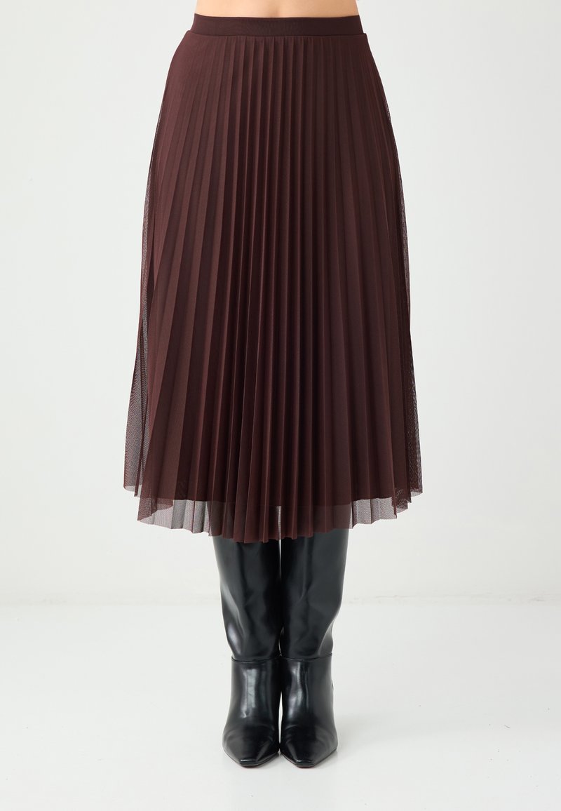 Jupe plissée marron en tissu transparent, superposée à un jupon uni, associée à des bottes noires pointues montantes jusqu'aux genoux.