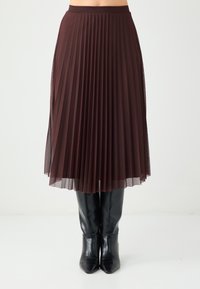 Jupe plissée marron en tissu transparent, superposée à un jupon uni, associée à des bottes noires pointues montantes jusqu'aux genoux.