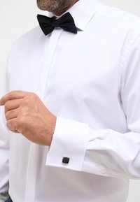 Weißes Hemd mit klassischem Kragen, schwarzer Fliege und schwarzen quadratischen Manschettenknöpfen. Glattes Material, sauber zugeschnitten mit Knopfverschluss.