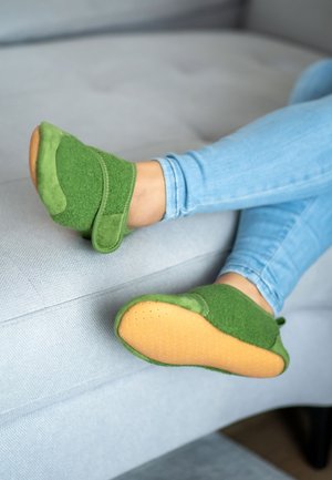 Grüne, gefilzte Hausschuhe mit einem sicheren Riemen verfügen über eine strukturierte Oberseite und eine glatte, sandfarbene Gummisohle, die auf einem hellgrauen Sofa resting.