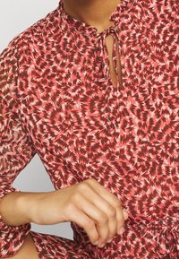 Hedvábná blůza červené a růžové barvy s potiskem, s průstřihovým výstřihem, detailem na zavázání a dlouhými rukávy. Lehká, texturovaná látka s jemným leskem.