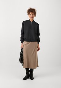 Melns zip-up bomberjakas ar ribotām aprocēm, apvienots ar beige satīna midi svārkiem. Melna zīmola somiņa ar auklu un augstiem melniem zābakiem.