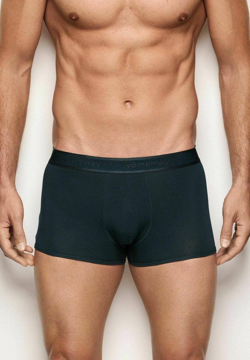 Torso masculino vistiendo boxers ajustados de color oscuro con el logo "yamamay" en la cinturilla, de pie frente a un fondo simple.