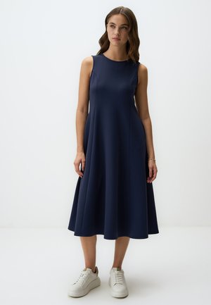 Navyblauwe mouwloze jurk met een uitlopende rok, gemaakt van gladde stof. Gepaard met witte sneakers voor een casual look.