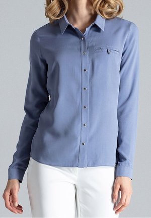 Femme portant une chemise bleu clair à manches longues avec une petite poche poitrine, associée à un pantalon blanc sur un fond gris uni.