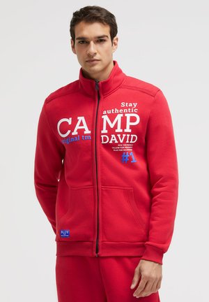 Junger Mann trägt ein rotes Sweatshirt mit Reißverschluss und weißem sowie blauem Schriftzug "Camp David" und "Stay authentic" auf der Vorderseite.