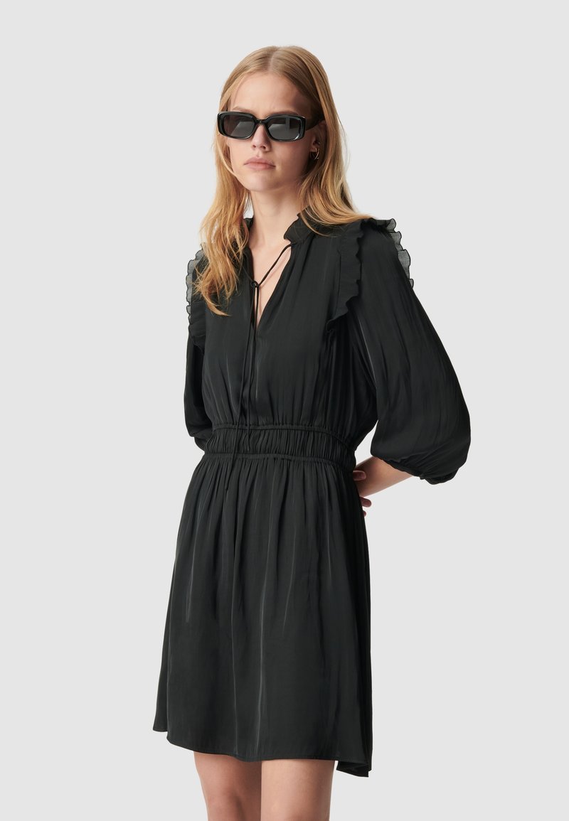 Abito nero con spalle arricciate, maniche larghe e vita elastica. Tessuto leggero e fluido che arriva a metà coscia, abbinato a occhiali da sole scuri.