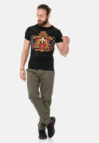 Schwarzes Grafik-T-Shirt mit einem lebendigen roten und goldenen Wappen-Design, kombiniert mit olivgrünen Hosen und schwarzen Schuhen.