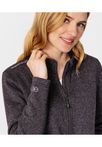 Chaqueta de forro polar gris con cremallera, fabricada en tejido texturizado, con cuello alto y cremallera negra. Presenta un logo en el hombro y detalles bordados en la manga.
