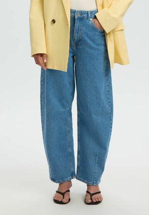 Femme portant un jean large bleu, un blazer jaune, un top blanc et des sandales noires, debout avec une main dans la poche.