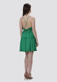 Un vestido verde con tirantes finos, cuerpo de encaje, diseño escalonado y dobladillo conVolante. La parte trasera presenta un escote bajo y tirantes cruzados.