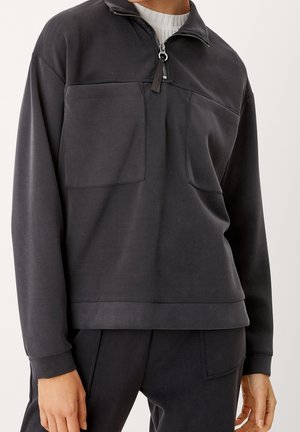 Sweatshirt noir à col zippé, avec deux poches avant et un tissu texturé, arborant une coupe décontractée et des poignets côtelés.