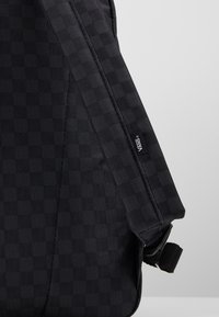 Sac à dos en tissu noir avec un motif à damier, une sangle réglable et une étiquette avec le logo Vans attachée sur le côté.