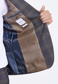 Blazer a quadri marrone e blu con fodera in raso beige, dotato di taschino e etichetta, che presenta un design su misura.